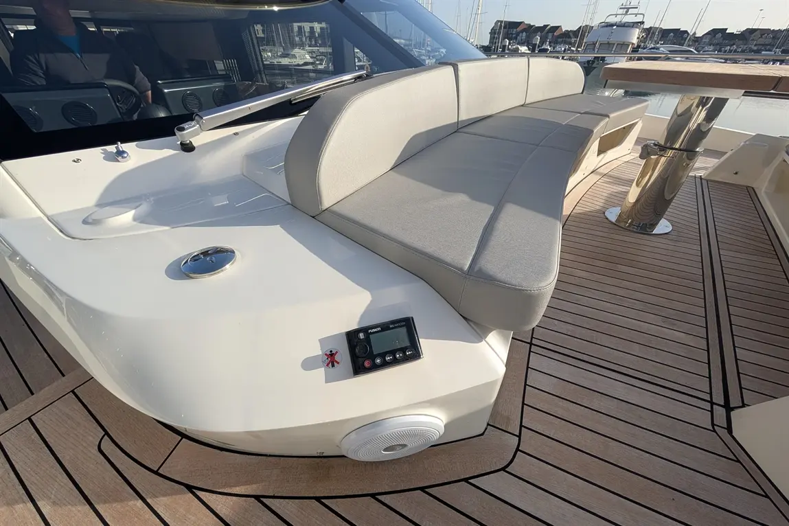 A modern boat's deck features a grey cushioned seating area, wooden flooring, a control panel, and a speaker.